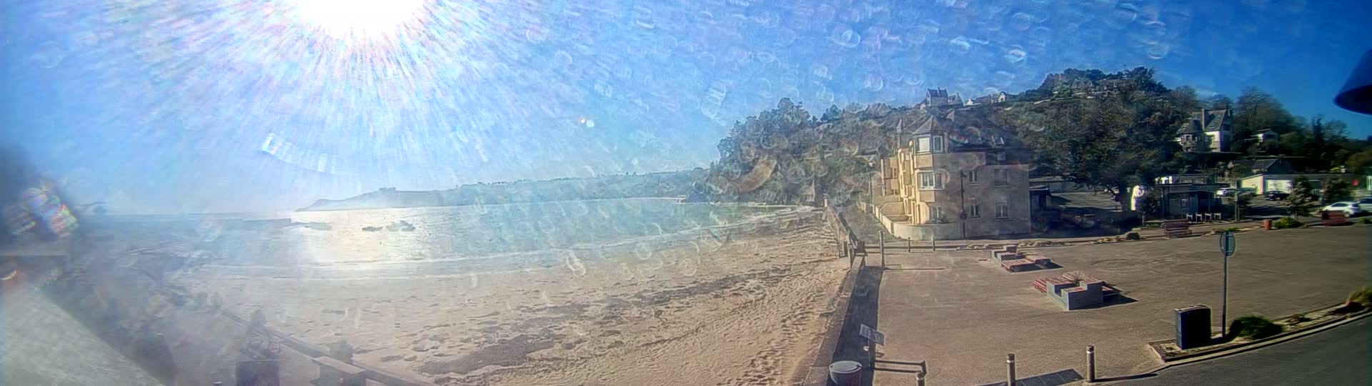 A sandy beach stretches alongside a calm, sunlit ocean, bordered by buildings and lush greenery on a bright, clear day.