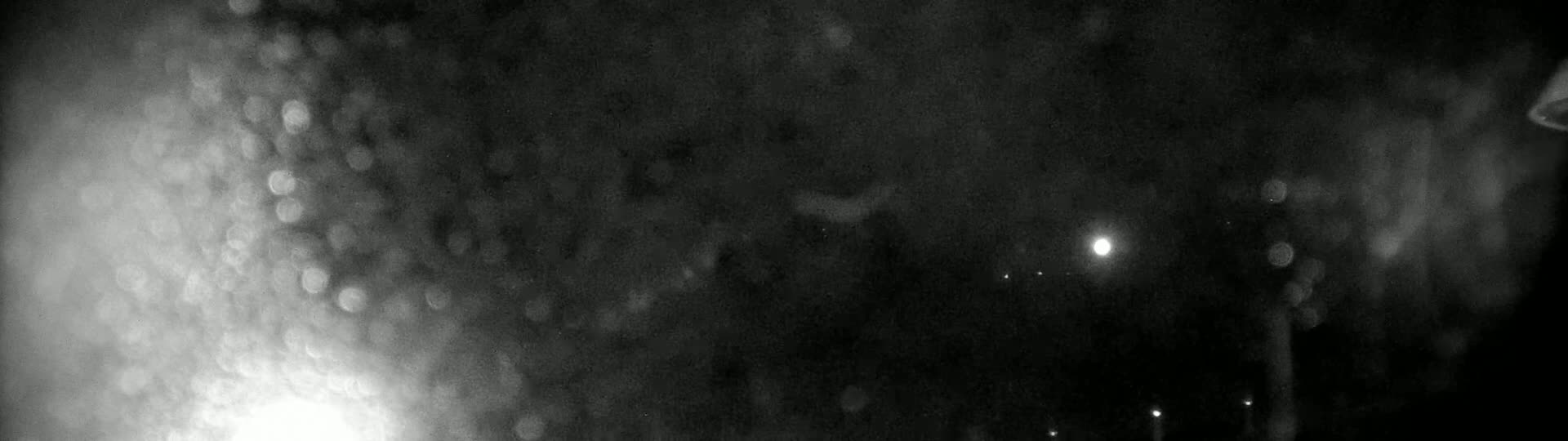 A blurry, nighttime image shows out-of-focus lights and what appears to be rain or snow falling on a dark surface.
