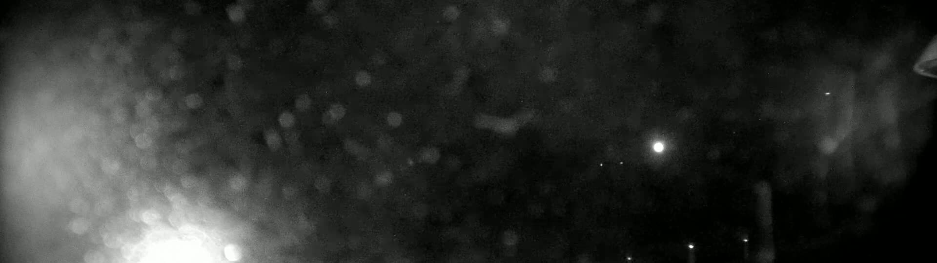A blurry, dark nighttime scene shows a heavy snow or rain falling in front of a light source.