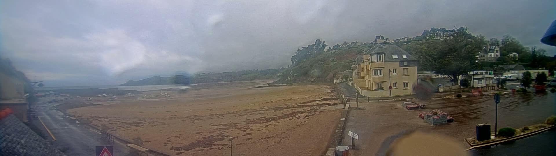 A sandy beach stretches out before a seaside town under an overcast sky, with rain-slicked roads and parking lots in the foreground.