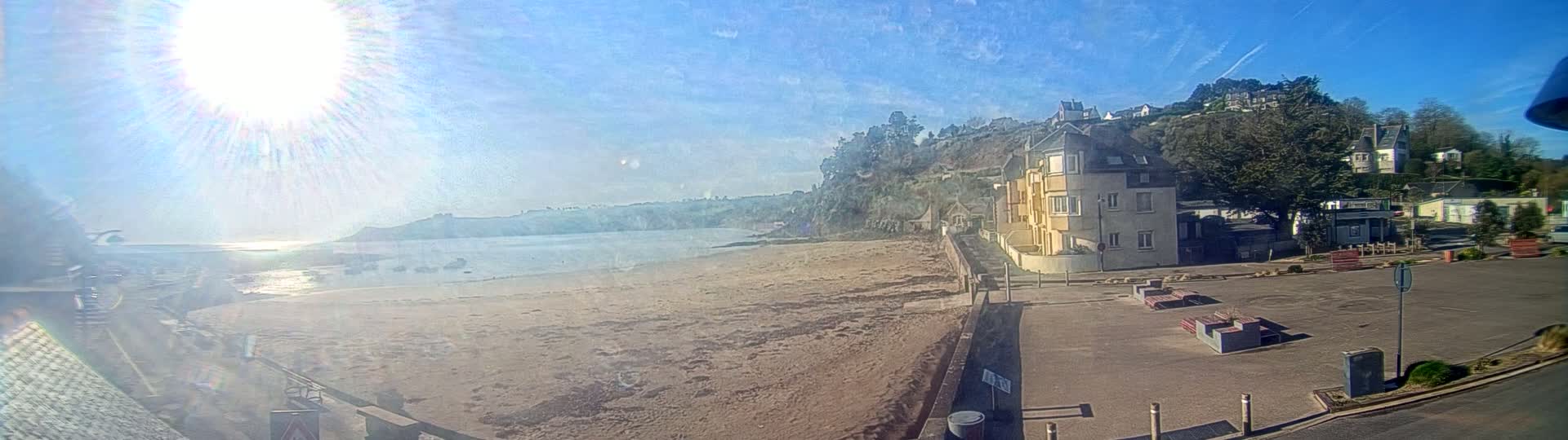 A sandy beach stretches out to a calm body of water under a bright, sunny sky, with buildings and greenery rising up on a hillside behind it.