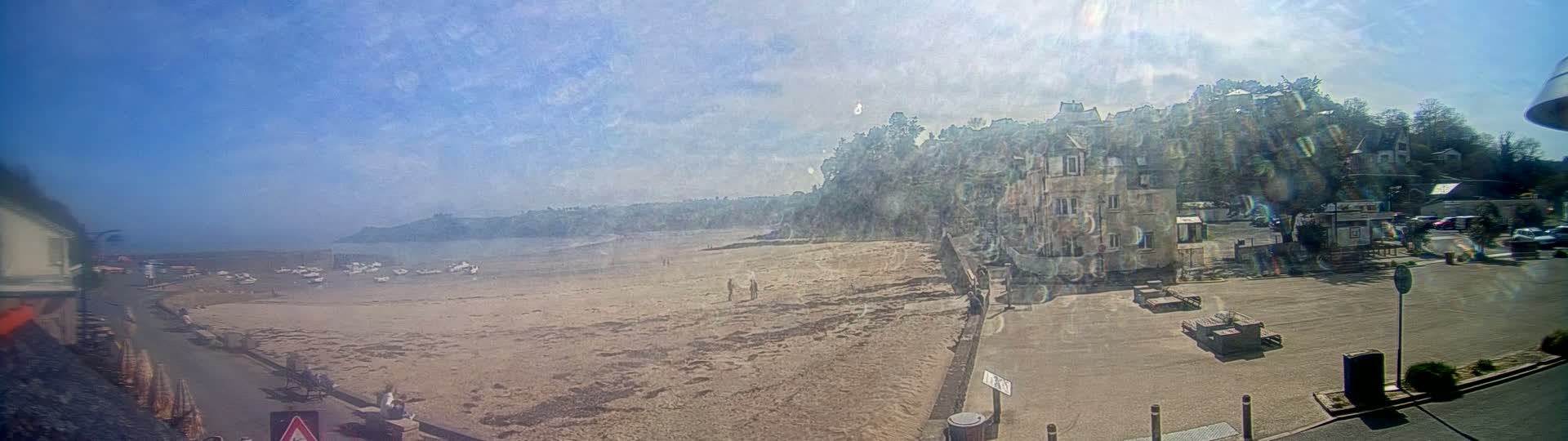 A sandy beach with several small boats on the water is visible below a hillside town under a mostly sunny sky.