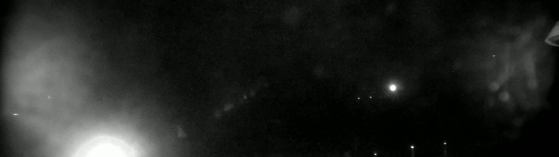 A nighttime outdoor scene shows a hazy, bright light source on the left and several fainter lights in the distance under a dark, cloudy sky.