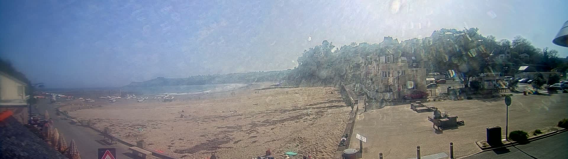 A sandy beach with a few people on it stretches out to a calm ocean under a bright, sunny sky, with buildings and trees rising on a hill behind it.