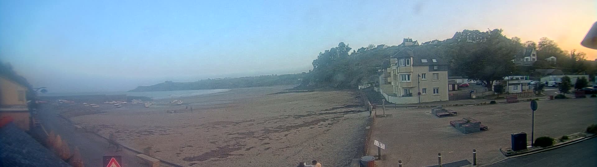 A hazy, pale-blue sky overlooks a sandy beach with several boats, a few people, and a building situated near a hillside with trees and more buildings in the distance.