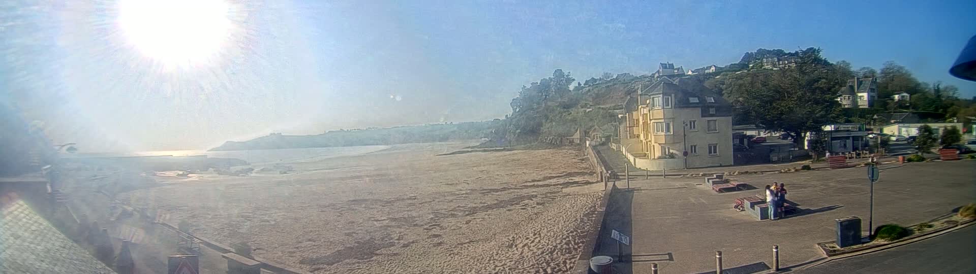 A sandy beach stretches out to a calm ocean under a bright, sunny sky, with buildings and a parking lot visible on the shore.