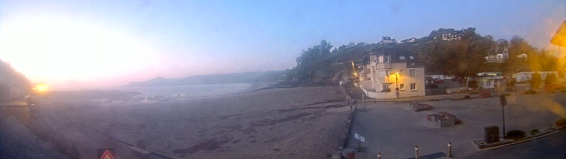 A hazy sunrise illuminates a sandy beach and waterfront buildings, with a hill covered in trees in the background.