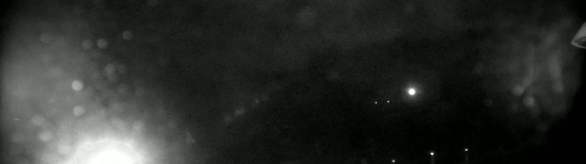 A blurry, nighttime image shows a bright light source in the lower left corner and several smaller, distant lights against a dark, hazy sky.