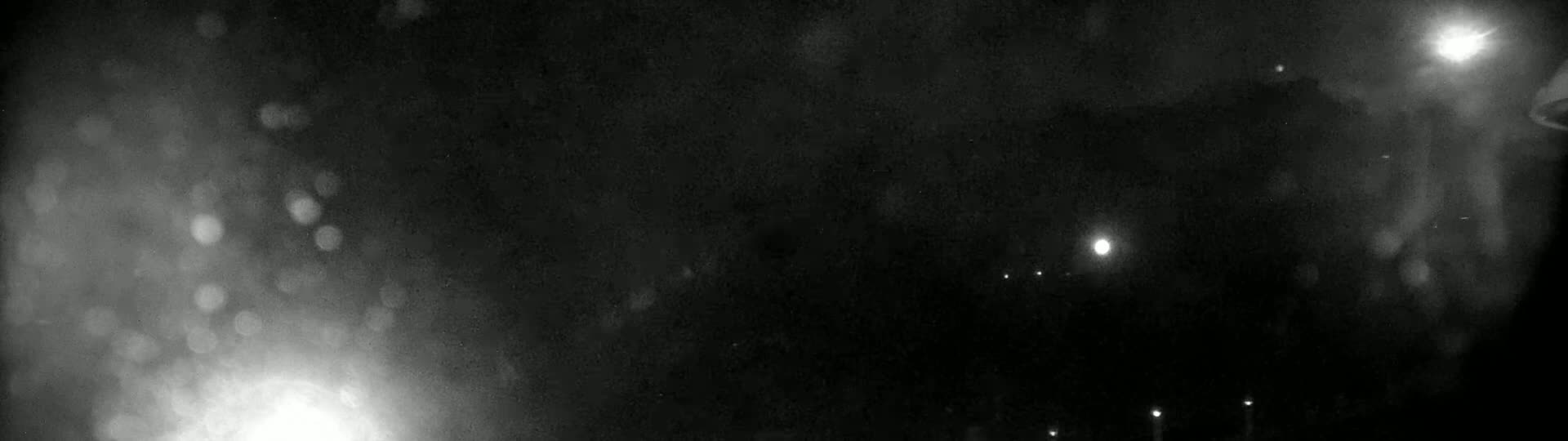 A nighttime, blurry, outdoor scene shows bright lights and indistinct shapes in the distance, possibly caused by rain or snow.