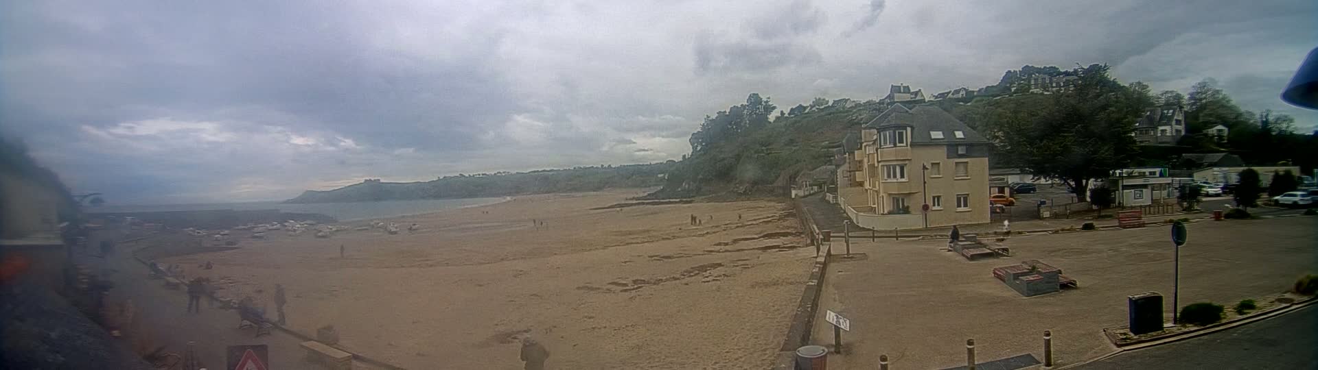 A sandy beach stretches out to a calm sea under a cloudy sky, with a few people scattered across the sand and a building visible on a nearby hillside.