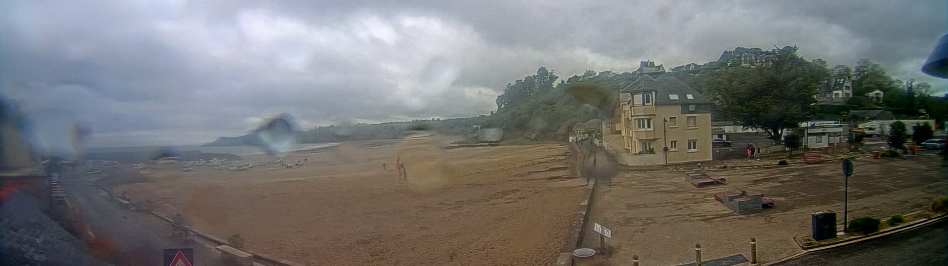 A rain-swept sandy beach with a few boats and a building are visible, with a hilly, tree-covered area in the background under an overcast sky.