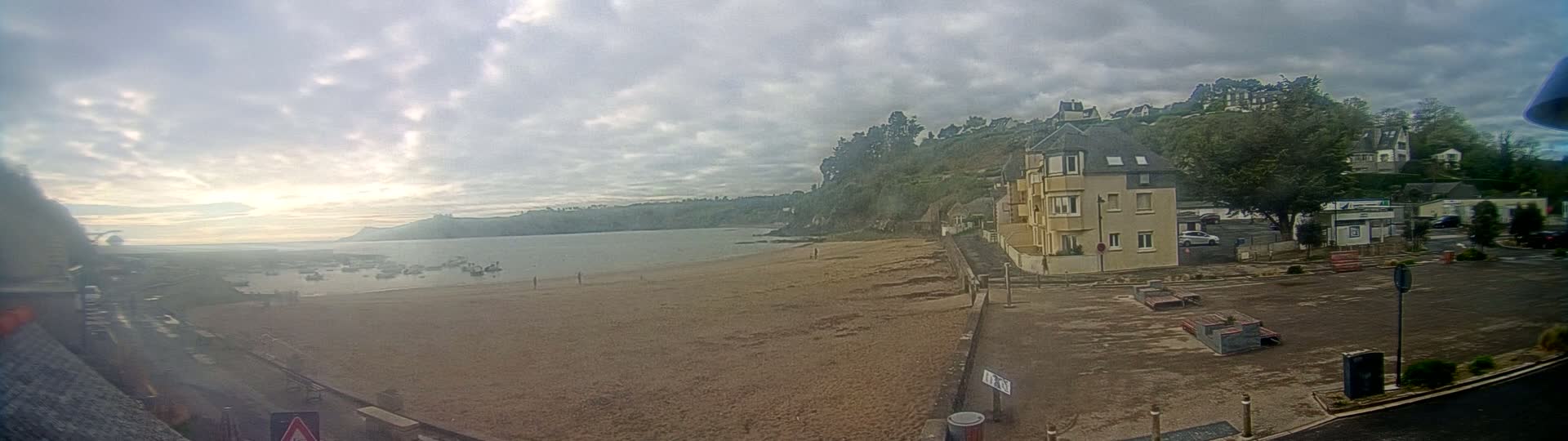 A sandy beach stretches out to a calm body of water under a mostly cloudy sky, with buildings and greenery visible on the far side of the beach.