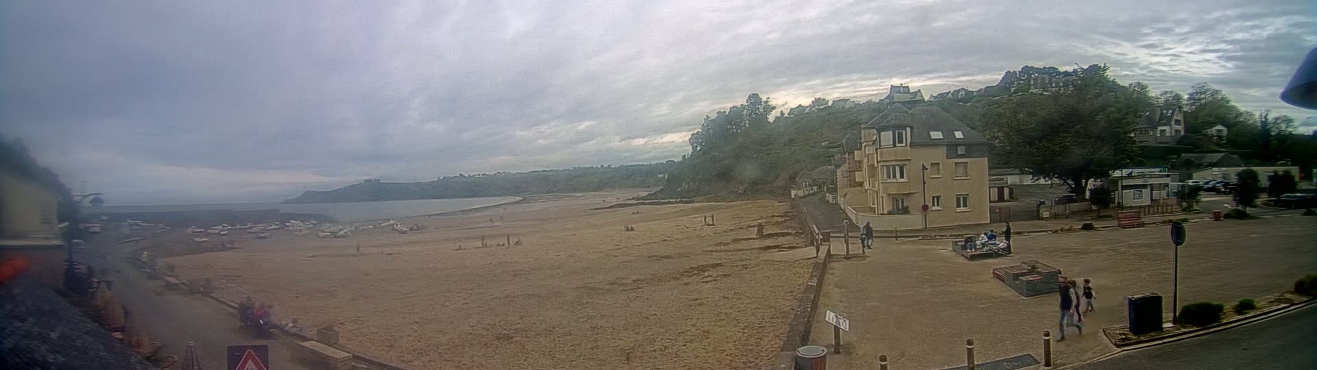 A sandy beach with a few people on it stretches out to a calm sea under an overcast sky, with buildings and trees situated on a hill overlooking the beach.