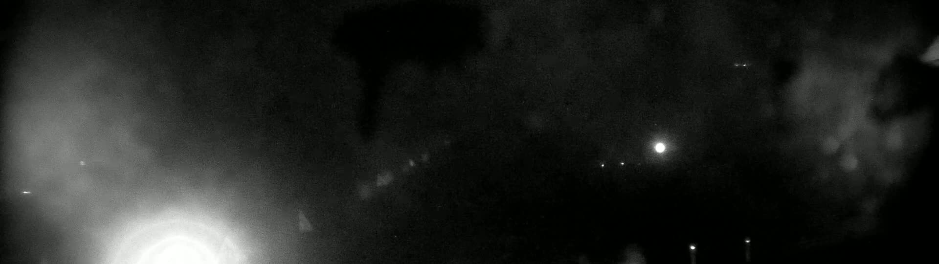 A nighttime scene shows a bright light in the lower left, several dim lights in the distance, and a hazy or cloudy sky.