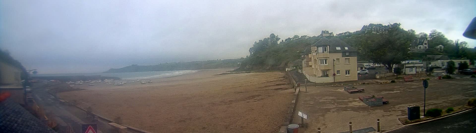 A sandy beach with several boats on the shore is seen below a residential area on a foggy day.