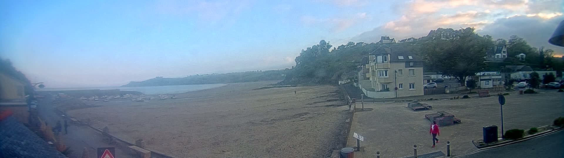 A hazy, overcast day reveals a sandy beach with several boats at low tide, a waterfront building, and a few people walking near parked cars on a nearby street.