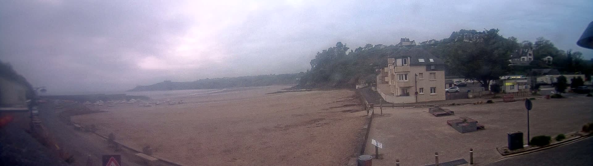 A sandy beach stretches out under an overcast sky, with a beige apartment building and parked cars visible nearby.