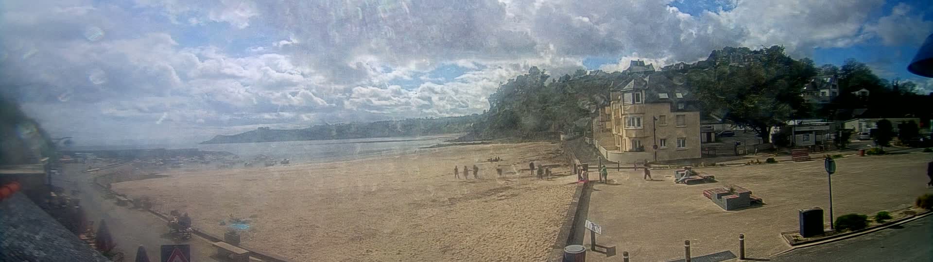 A sandy beach with a few people on it is visible beneath a partly cloudy sky, next to a town with buildings on a hillside.