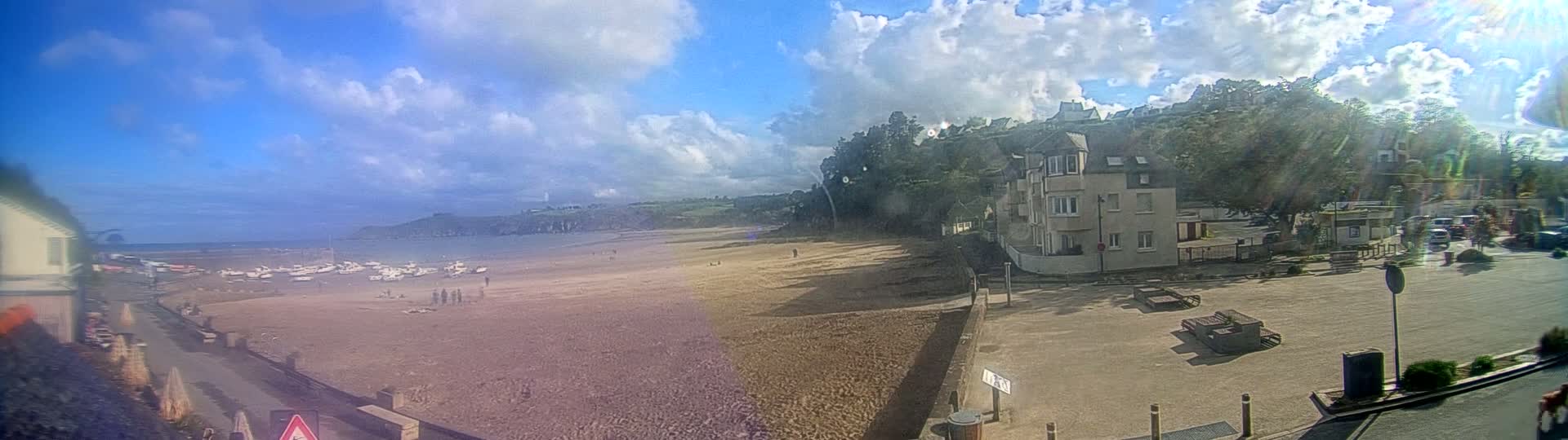 A sandy beach with boats on the water, a few people, and buildings on a hillside under a partly cloudy sky.