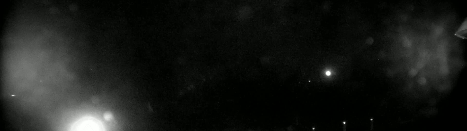 A nighttime, blurry outdoor scene shows a bright light and several fainter lights, possibly from distant sources, under a dark, possibly snowy or dusty sky.