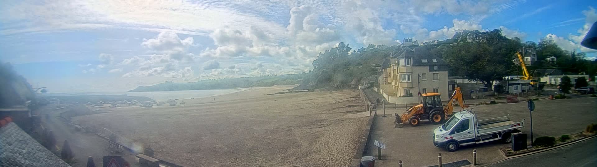 A sandy beach stretches out to a calm bay under a partly cloudy sky, with a small town, construction equipment, and a van parked nearby.