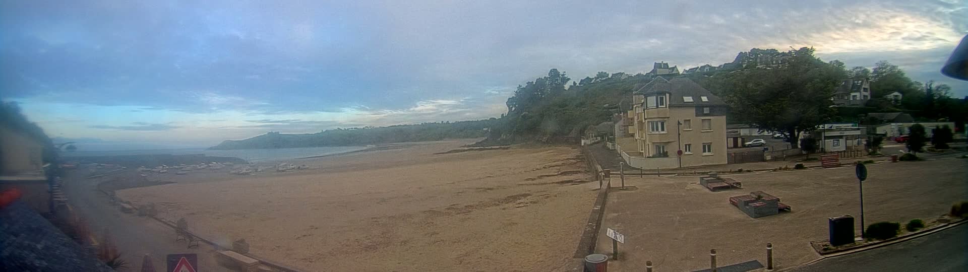 A sandy beach stretches out to a calm body of water under a mostly cloudy sky, with a multi-story building and green hillside visible in the background.