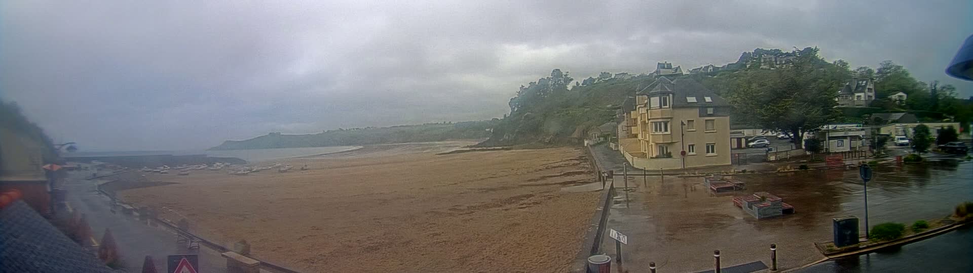 A sandy beach with boats on the water is seen from a waterfront vantage point on an overcast day, with rain-slicked streets and buildings visible beyond a seawall.