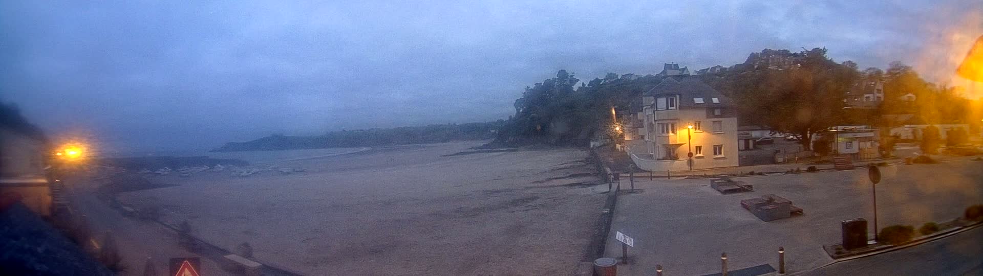 A sandy beach stretches out to a calm, dusky ocean under an overcast sky, with a building and street scene visible in the foreground.