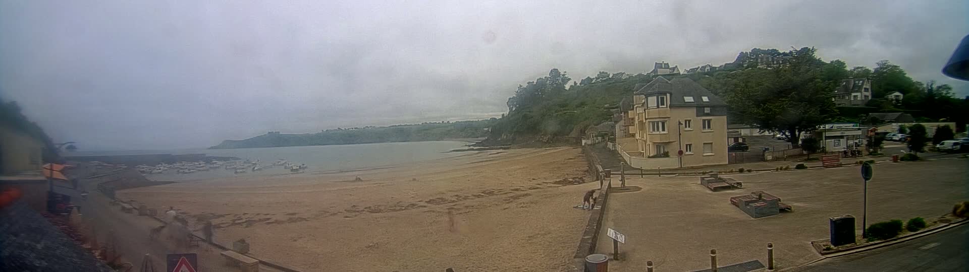 A sandy beach with several small boats in the water is visible under an overcast sky, next to a parking lot and a residential building on a hillside covered in green trees.