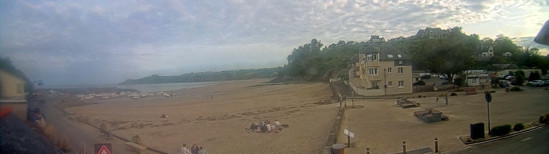 A sandy beach with several boats pulled ashore is visible beneath a cloudy sky, with a residential area and parking lot overlooking it.