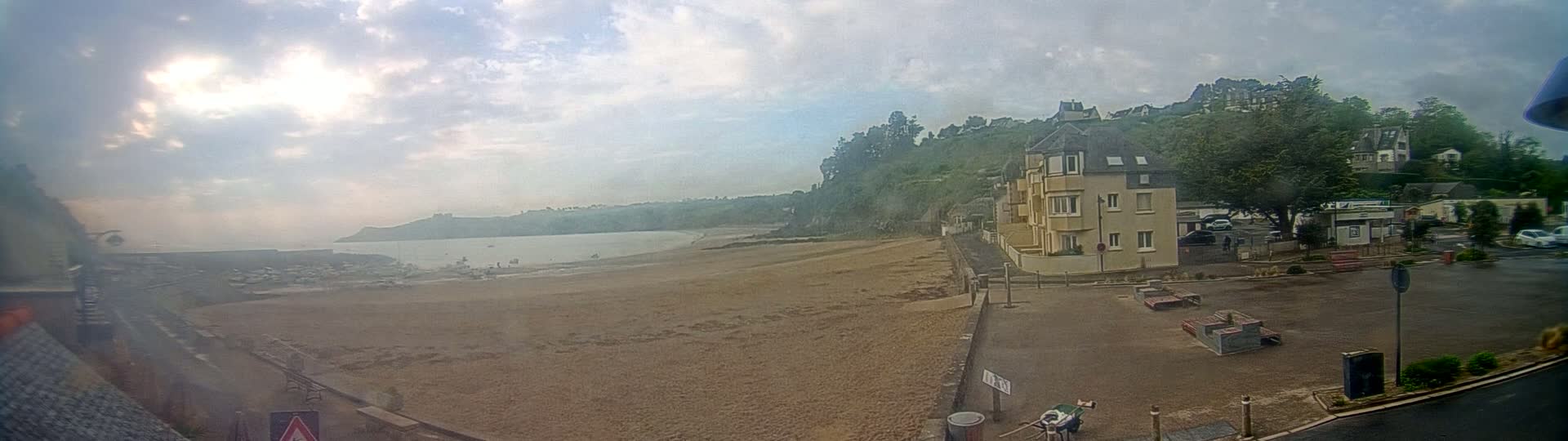 A sandy beach stretches along a calm ocean under a partly cloudy sky, with buildings and green hills rising behind it.
