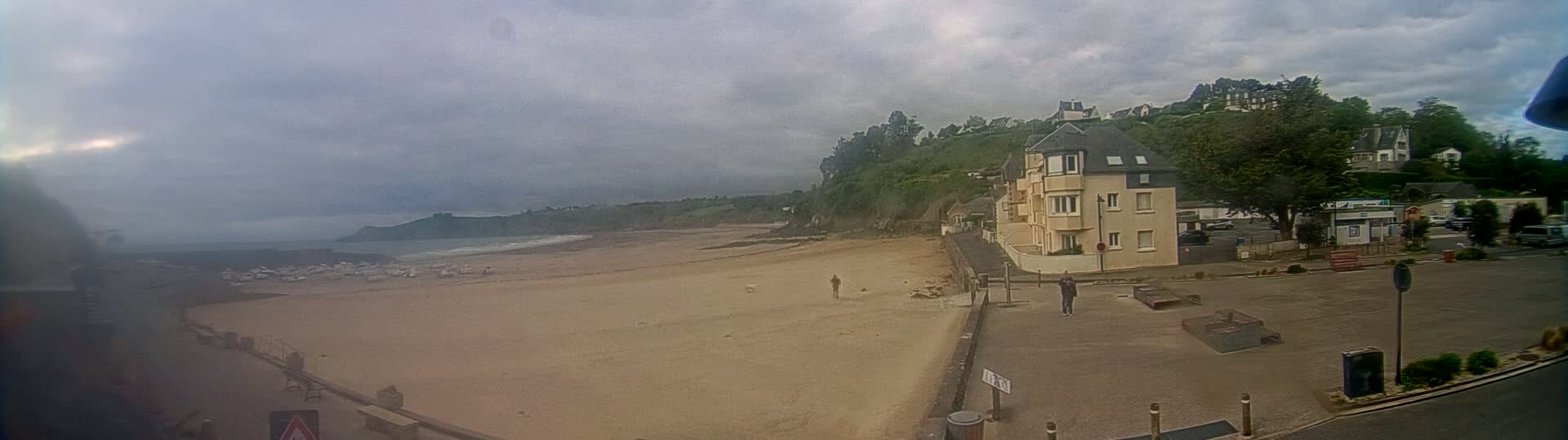 A sandy beach stretches out under a cloudy sky, with buildings and a parking lot visible in the foreground.