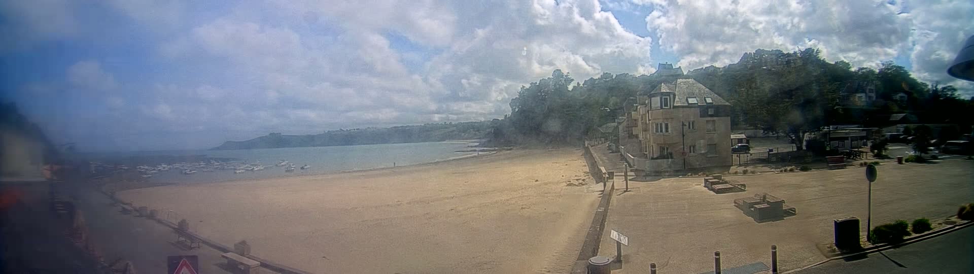A sandy beach with boats in the water and a building on a hill under a partly cloudy sky.