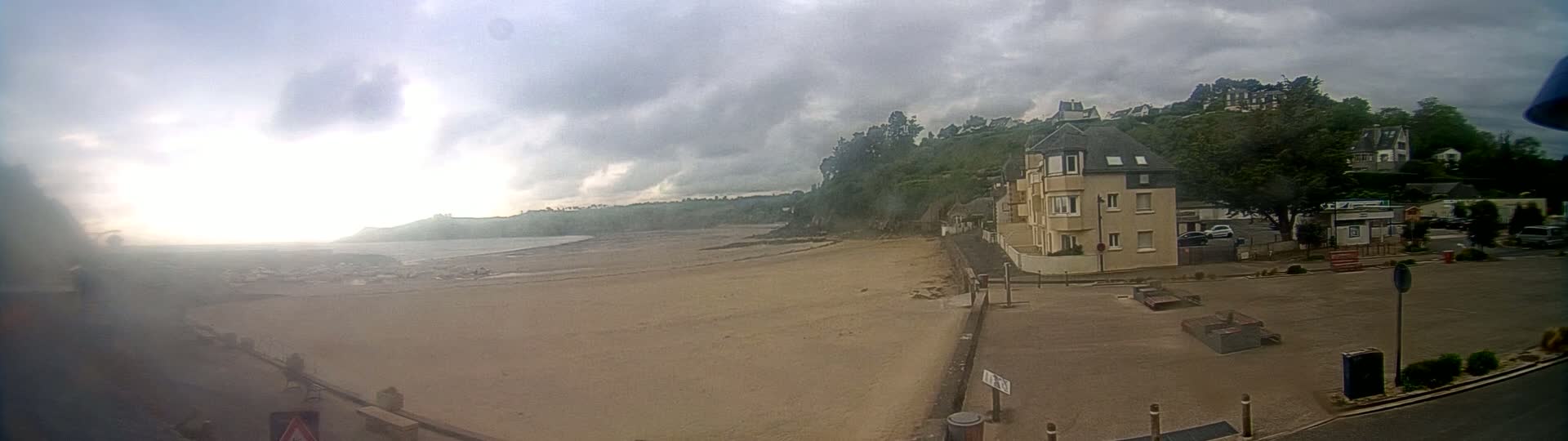 A sandy beach stretches out to a calm body of water under a cloudy sky, with a multi-story building and other structures situated on a hill overlooking the beach.