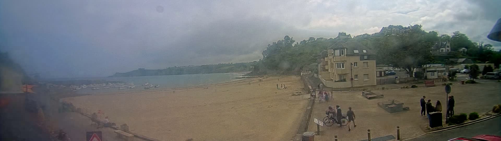 A sandy beach with a calm body of water and several boats is overlooked by a multi-story building and a hill covered in green vegetation under an overcast sky.