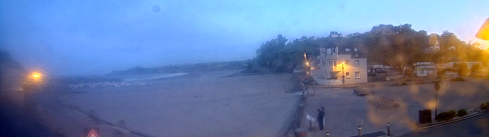 A hazy, dusky view of a beach with a building and trees behind it, under a pale, almost blue sky.