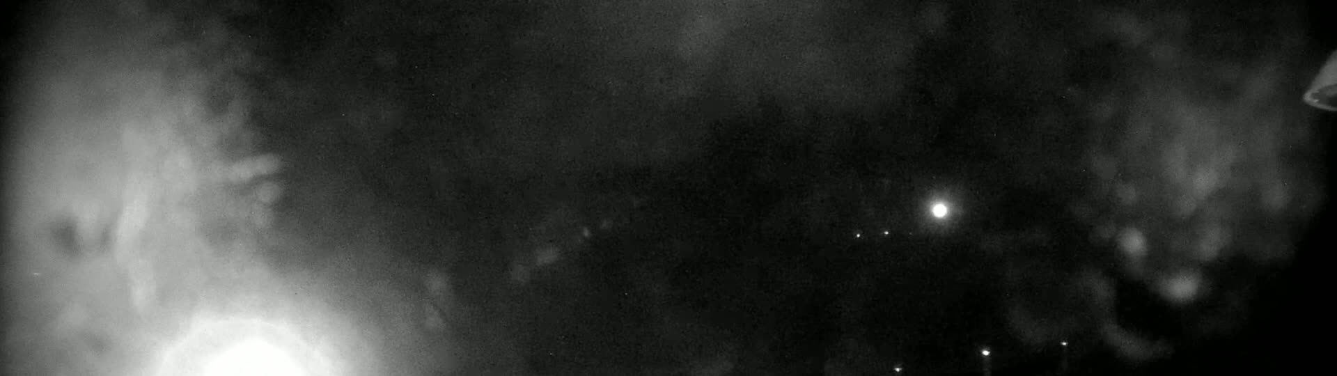 A nighttime, blurry view shows a bright light on the lower left, possibly a fire, and several fainter lights in the distance, under a hazy, dark sky.