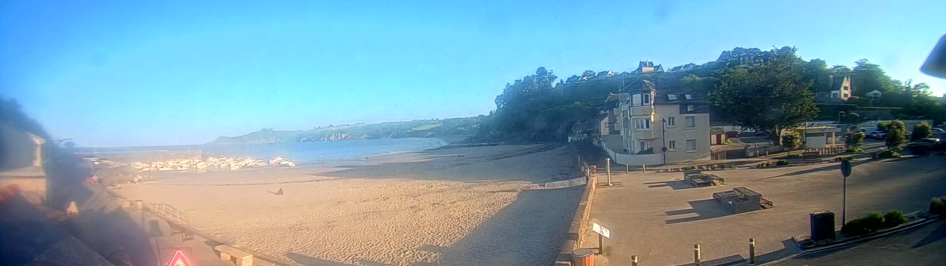 A sandy beach with numerous boats on the shore is adjacent to a town with buildings on a hillside, all under a clear blue sky.