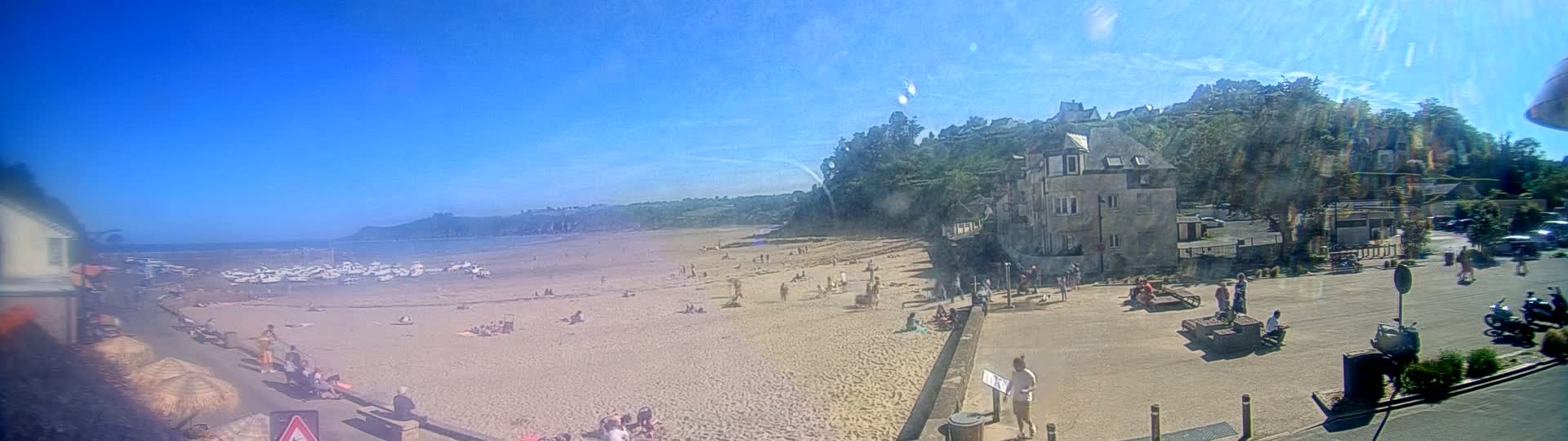 A sunny day at a sandy beach, with many people relaxing and swimming, boats on the water, and buildings on a hill overlooking the scene.