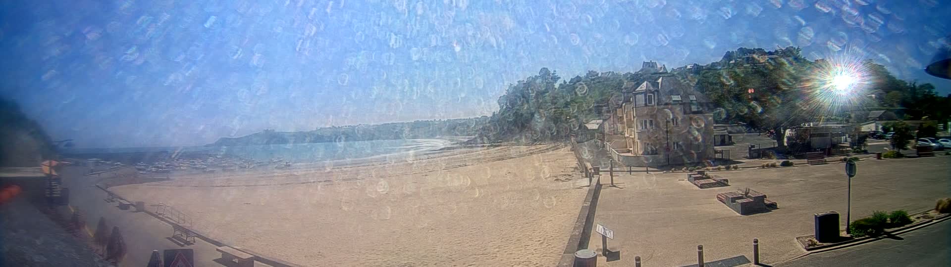 A sandy beach stretches along a calm ocean under a bright, sunny sky, with buildings and green hills rising in the background.