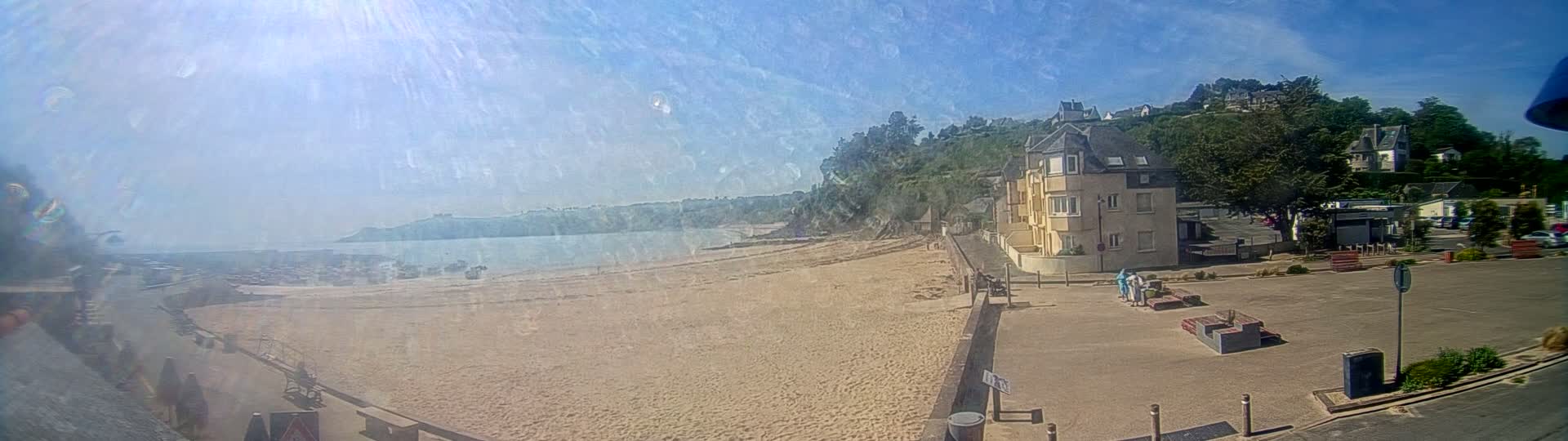 A sandy beach stretches toward a calm ocean under a bright, sunny sky, with a building and parking lot situated nearby on a hill overlooking the scene.
