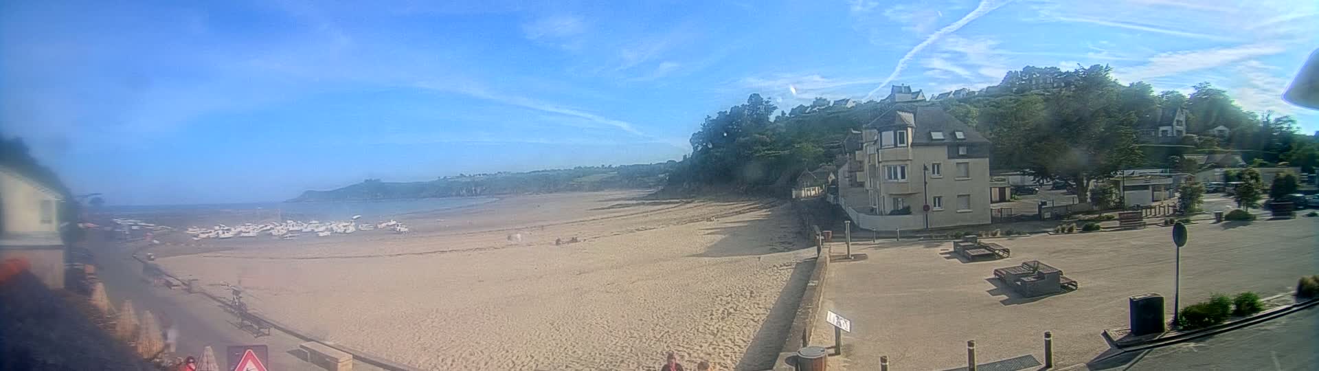 A sandy beach stretches out to a calm ocean under a mostly clear, sunny sky, with a few buildings and green vegetation visible on the adjacent hillside.