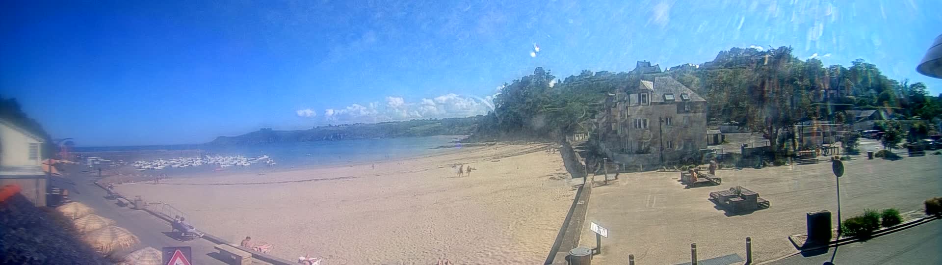 A sandy beach with numerous boats in the water is viewed from a hilltop overlooking buildings and a parking lot on a sunny day with some clouds.