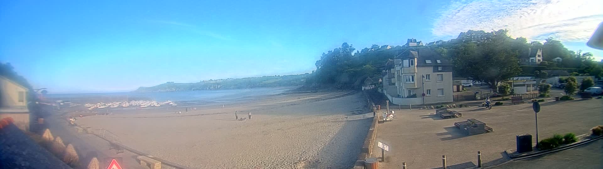 A sandy beach with numerous small boats on the water stretches out beneath a clear blue sky, bordered by a hillside with buildings and lush green vegetation.