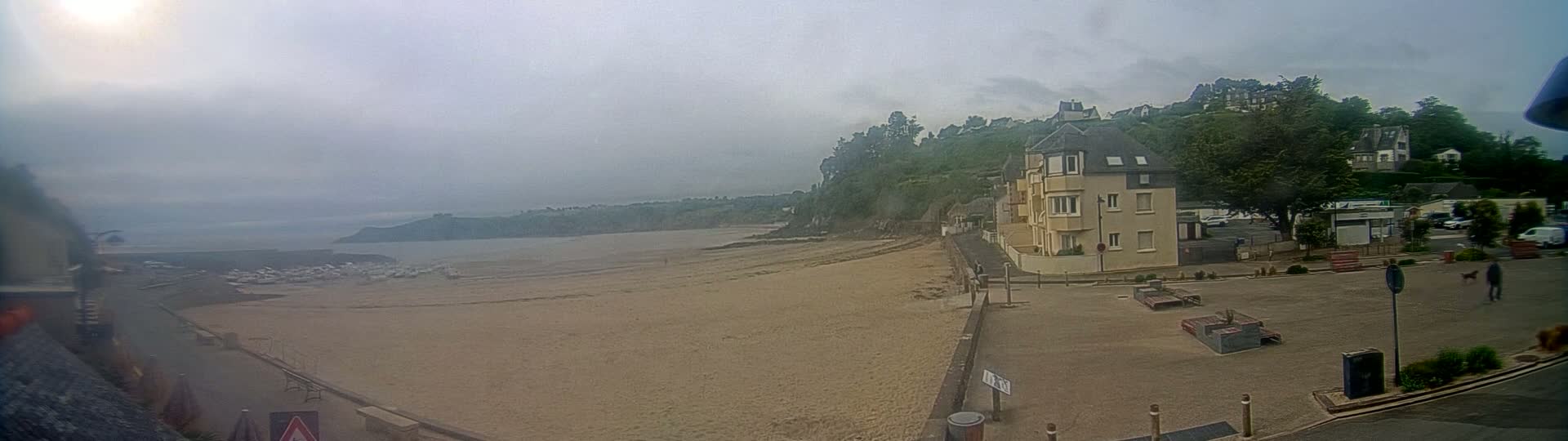 A sandy beach stretches out to a calm, overcast ocean under a hazy sky, bordered by a seaside town with buildings and green hills.