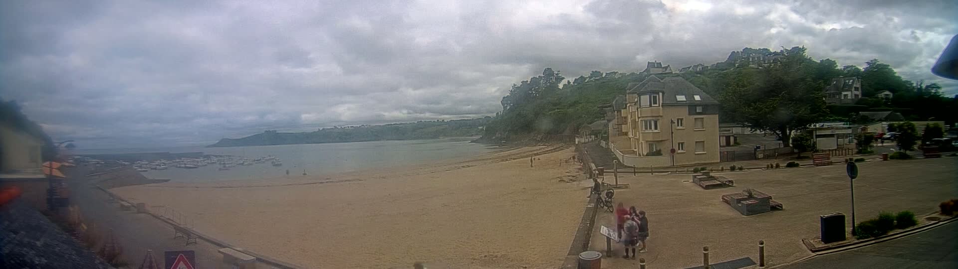 A sandy beach with a calm body of water, boats, and a few people are visible under a cloudy sky, backed by buildings and lush green hills.