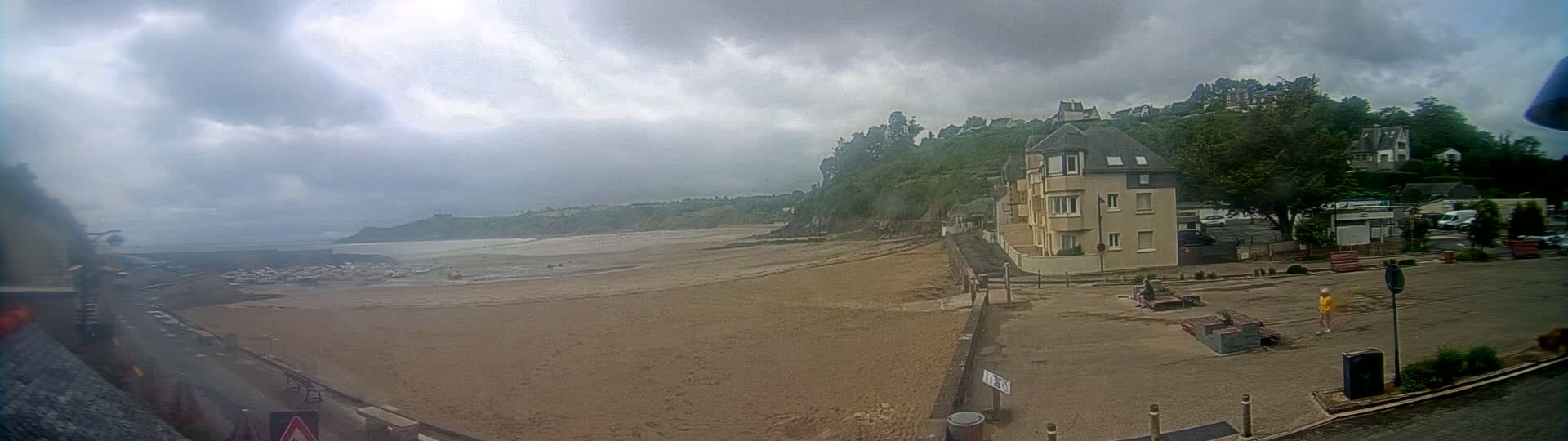 A sandy beach stretches out to a calm sea under a cloudy sky, with a residential building overlooking the scene.