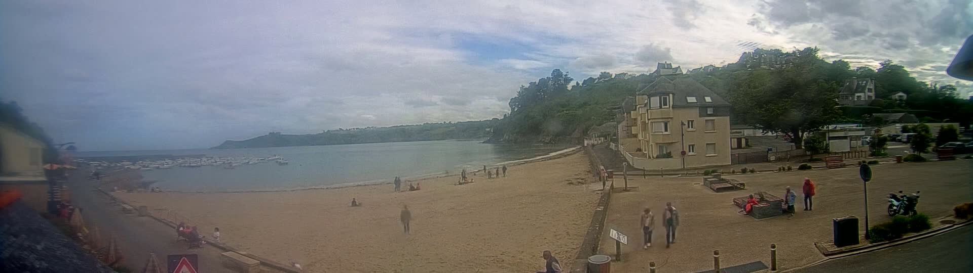 A sandy beach with several people on it is situated next to a calm bay with boats, overlooked by buildings and green hills under a cloudy sky.