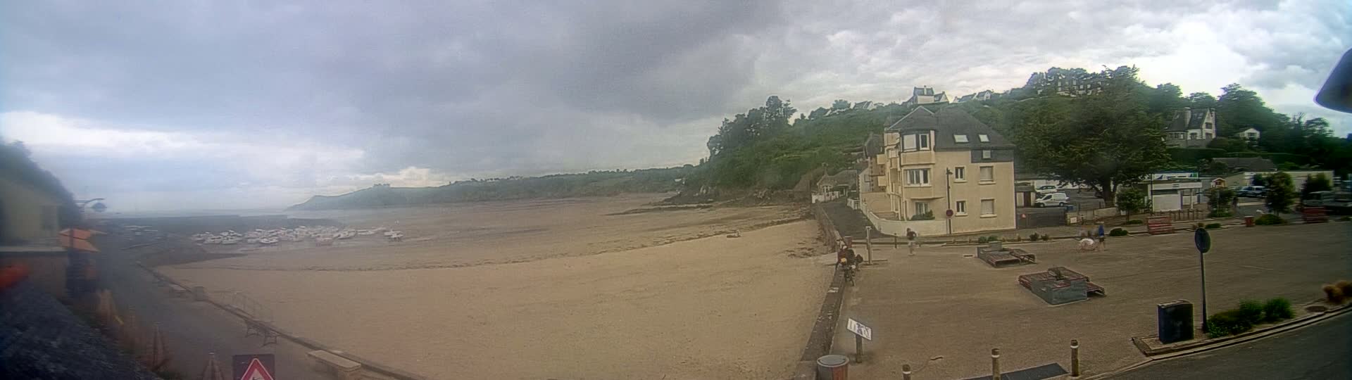 A sandy beach stretches out under an overcast sky, with a multi-story building and several smaller structures situated on a nearby hillside overlooking a parking lot and a small town.