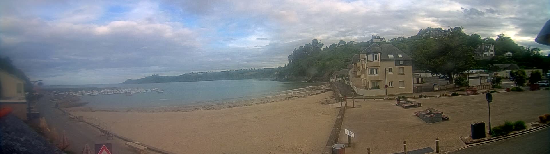 A sandy beach with boats in the water is next to a parking lot and buildings, under a cloudy sky.